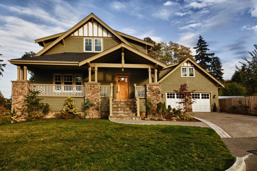 A Craftsman-style house with a covered porch, wooden front door, stone columns, dormer windows, attached garage, and a neatly manicured lawn under a partly cloudy sky features expert roofing Burke craftsmanship.