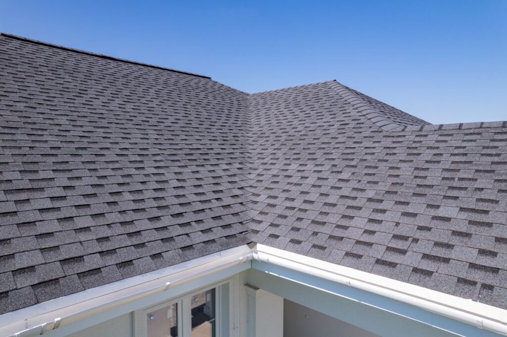 A close-up view of a house roof with dark gray asphalt shingles by roofing Springfield, featuring intersecting slopes and a white gutter along the edge, set against a clear blue sky.