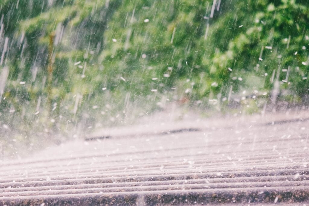 Heavy rain falls on a slanted, ridged metal roof from Roofing Alexandria with blurred green trees in the background, creating a sense of motion and wet, stormy weather.