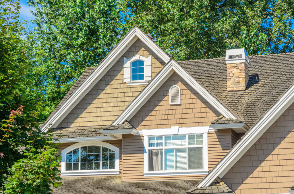 The upper part of a house with brown shingle siding, white trim, multiple gabled roofs, and several windows showcases expert roofing McLean VA style, surrounded by green leafy trees under a clear sky.