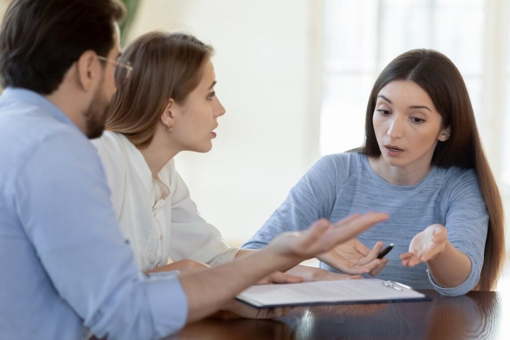 Three people sit at a table having a serious discussion about roofing in Alexandria. A woman in a blue shirt gestures with her pen, speaking to a man and woman who listen closely, with documents and a clipboard on the table.