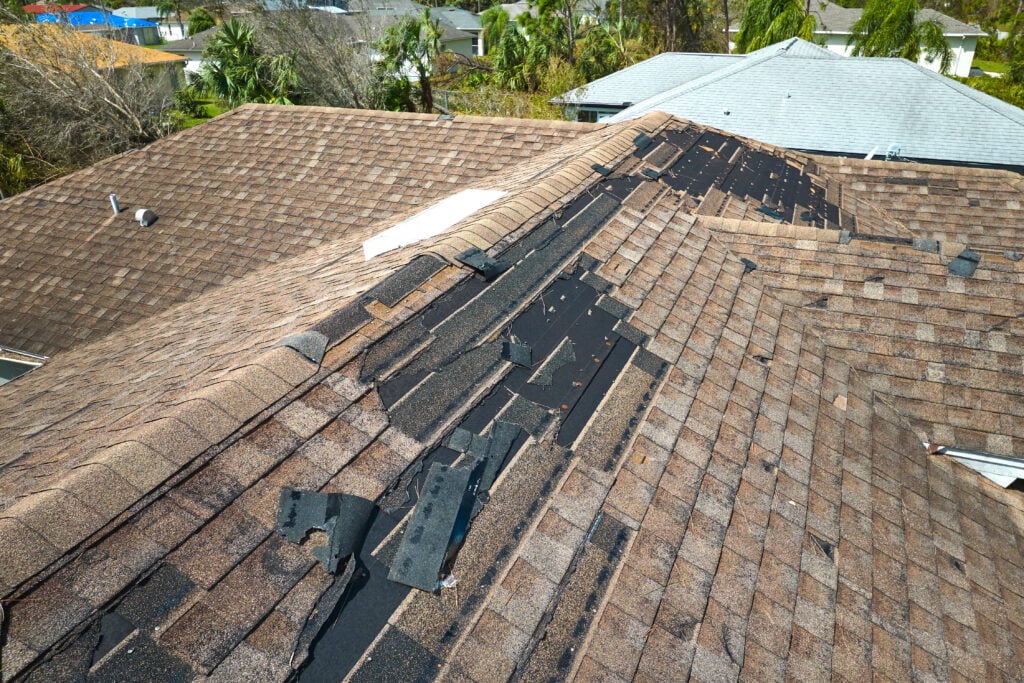 A house roof in Burke with numerous missing and damaged shingles exposes the black underlayment beneath. The damage runs along the ridge, likely caused by strong winds or a recent storm. Neighboring roofs remain intact.