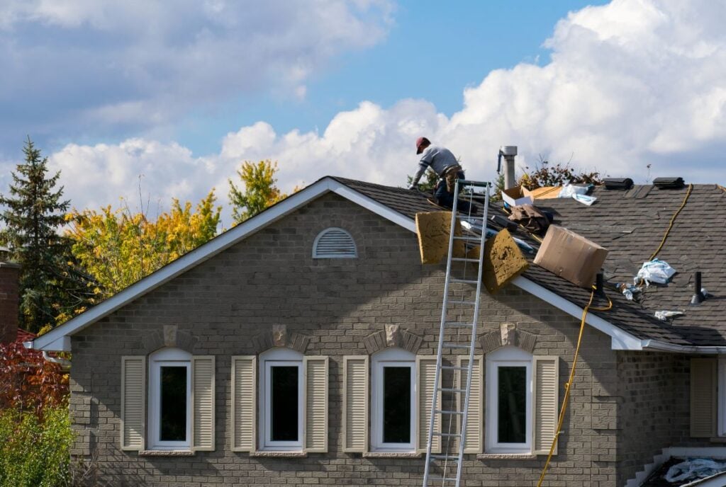 A worker wearing safety gear repairs the roof of a house using tools and materials for a roofing Mount Vernon project, with a ladder propped against the roof and boxes nearby. Trees with autumn foliage are in the background under a cloudy sky.