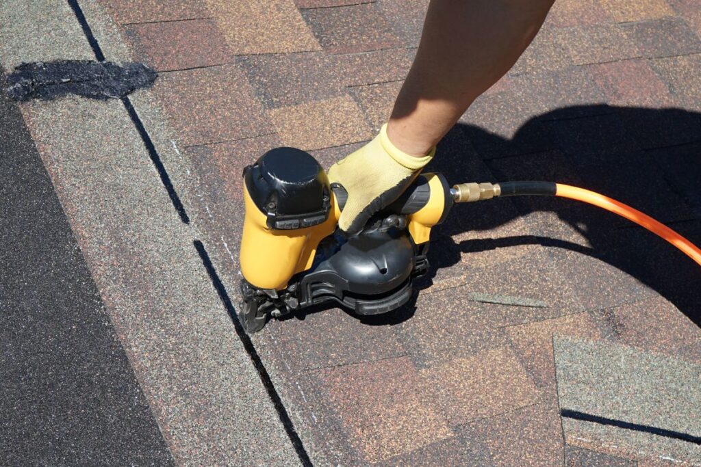 A person wearing a yellow glove uses a pneumatic nail gun to secure asphalt shingles on a house roof in roofing Springfield. The shingles are brown, and the nail gun is connected to an orange air hose.