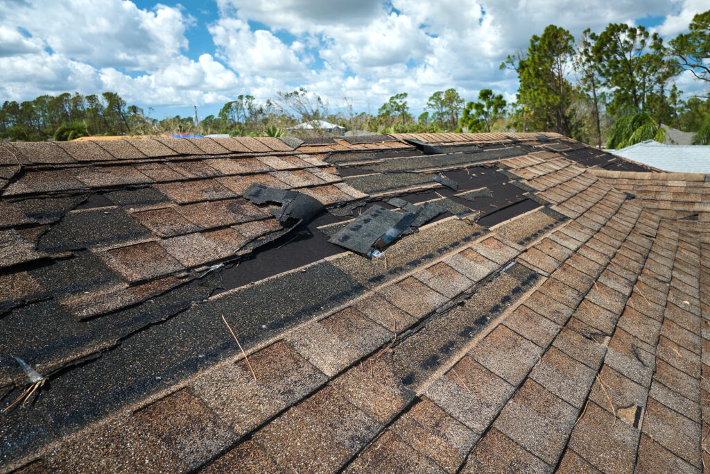 A roof in McLean, VA with brown asphalt shingles shows storm damage—missing and displaced shingles expose black underlayment beneath. Roofing experts in McLean, VA recommend prompt repairs. Trees and a partly cloudy sky are visible in the background.