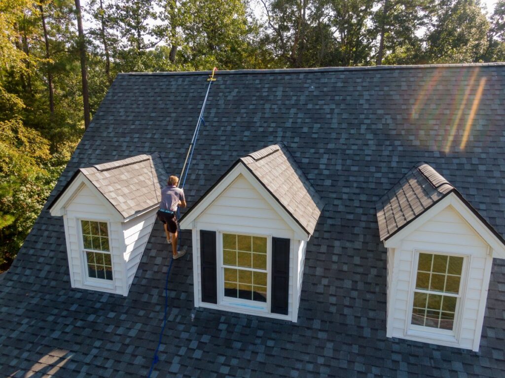 A person uses a long pole to clean the roof of a house with gray shingles and two dormer windows, surrounded by trees on a sunny day—typical of roofing in Mount Vernon.
