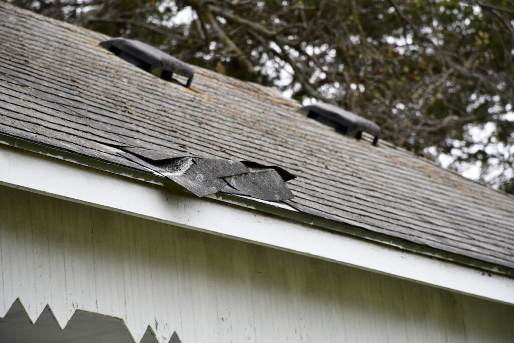 A house roof with several loose, lifted shingles near the edge, indicating possible storm or wind damage. Tree branches are visible in the background above the roofline—a sign it’s time for expert roofing McLean VA services.