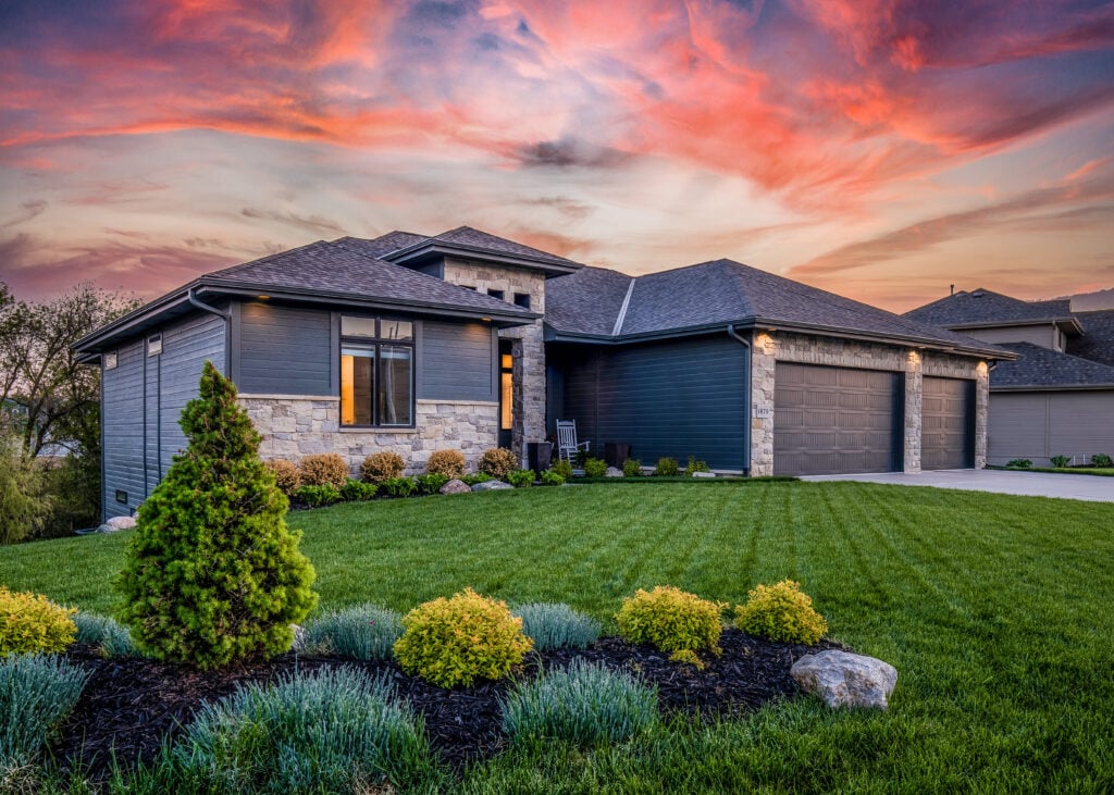 Modern single-story house with gray siding, stone accents, and expert roofing Fairfax Station style, features a well-manicured green lawn and vibrant landscaping. The sky glows with orange and pink hues at sunset.