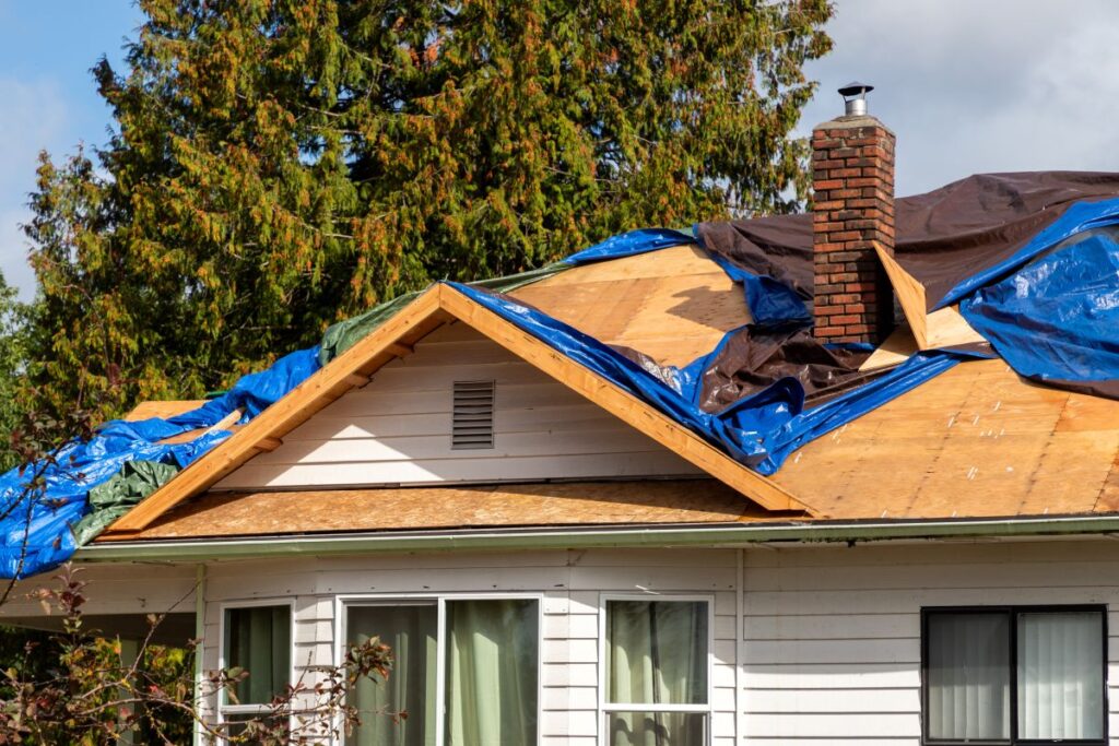 A house with part of its roof under repair, covered by blue and black tarps. Exposed wooden beams and a brick chimney are visible, with trees in the background—an example of roofing Springfield projects in progress.