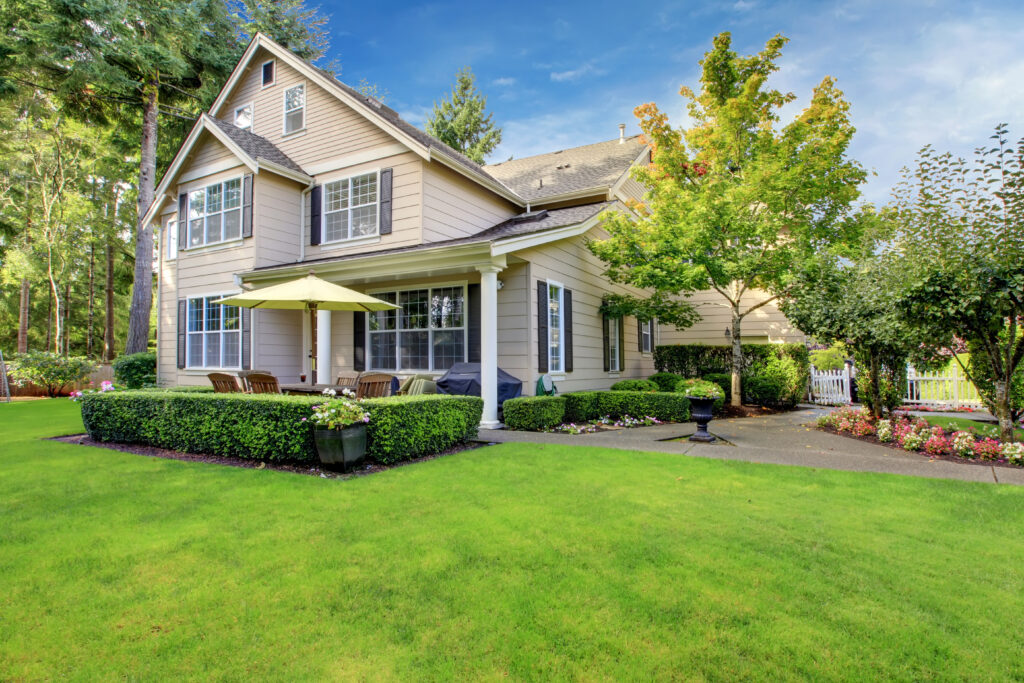 A two-story beige house with large windows and a well-maintained lawn in roofing Fairfax Station style. The patio area features outdoor seating and a yellow umbrella, surrounded by green bushes, trees, and bright flowerbeds.