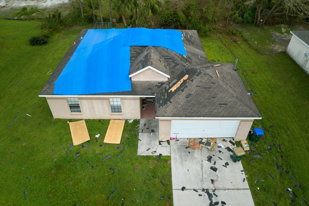 A house with roof damage covered by a large blue tarp, missing shingles, and debris scattered in the yard and driveway, suggesting recent storm or hurricane damage in need of expert roofing Burke services.