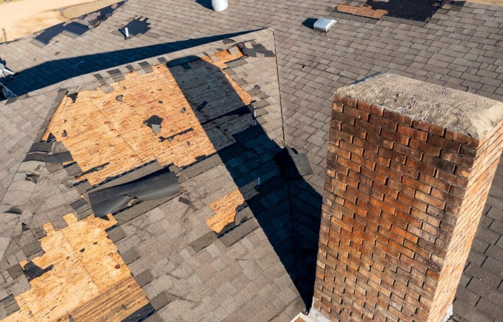 A damaged roof with missing and broken shingles, exposing plywood underneath near a large brick chimney. The roof in this Mount Vernon home shows signs of wear and significant storm damage, highlighting the need for expert roofing services.