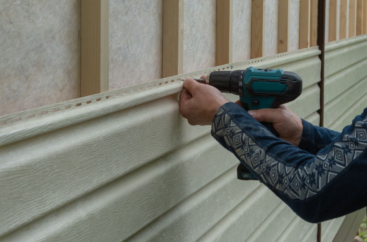 A person uses a power drill to install light-colored siding panels onto the outside wall of a building with wooden framing during roof repair in Lorton.