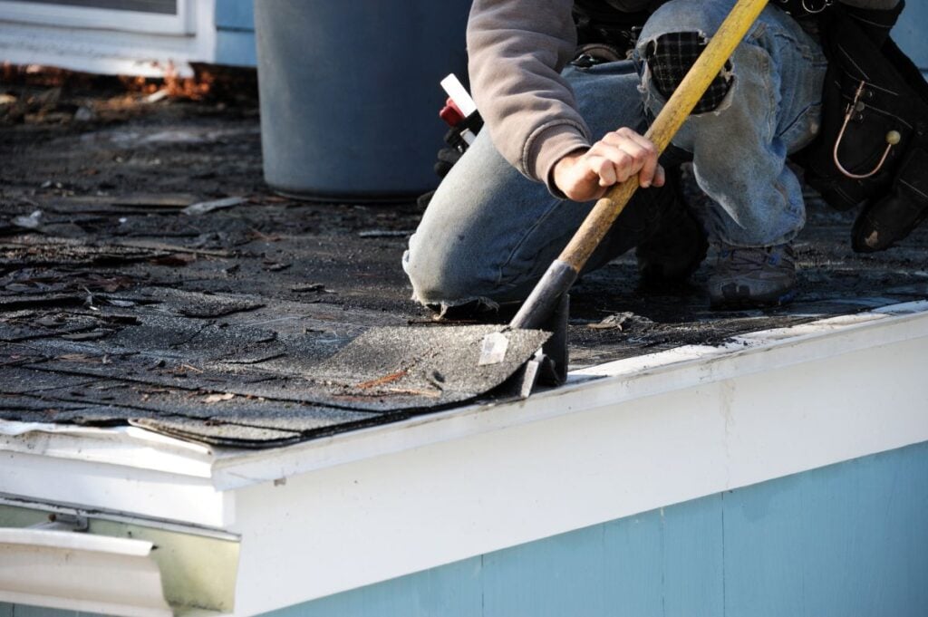 A person in jeans and a hoodie uses a shovel to remove old, damaged shingles from a roof during a roofing Mount Vernon repair or renovation project.