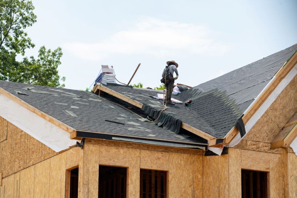 A roofing Springfield construction worker wearing a hat installs roofing materials on a partially completed house roof under a bright sky. Shingles and tools are scattered around the work area.