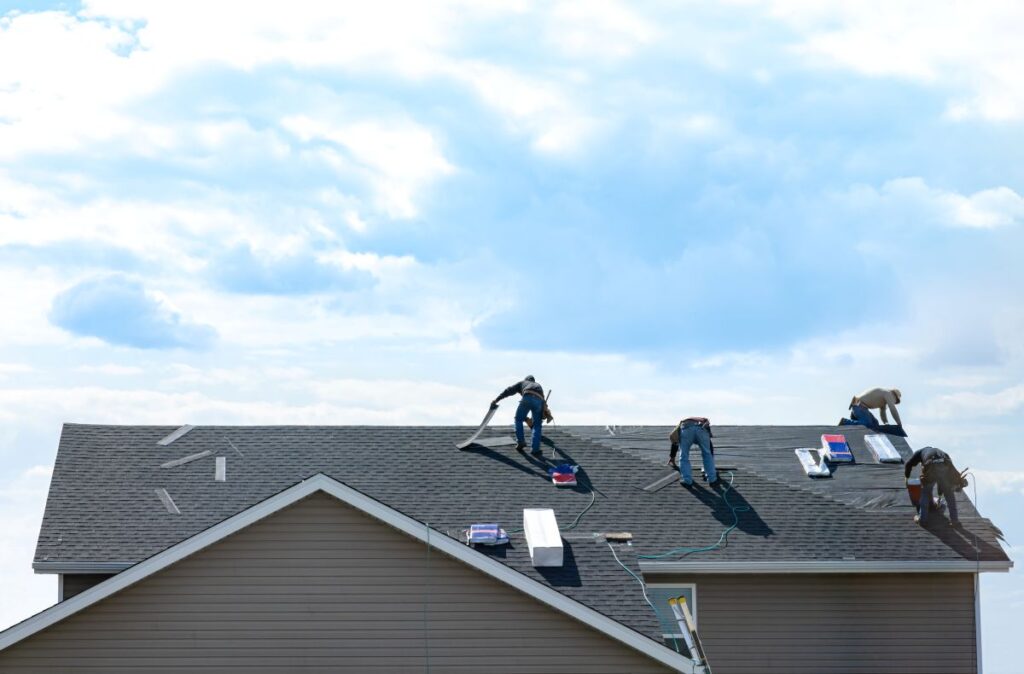 Four workers install or repair shingles on a house roof in Springfield, with tools and materials scattered nearby under a partly cloudy sky—showcasing expert roofing Springfield services in action.