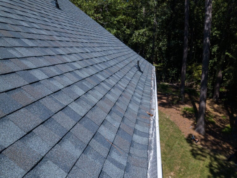 A close-up view of a sloped house roof with gray asphalt shingles—highlighting quality roof materials Falls Church residents trust—plus a white gutter, surrounded by trees and grass on a sunny day.