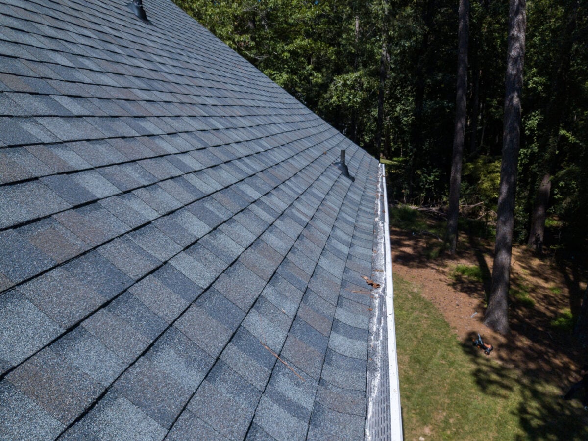 A close-up view of a sloped house roof with gray asphalt shingles—highlighting quality roof materials Falls Church residents trust—plus a white gutter, surrounded by trees and grass on a sunny day.