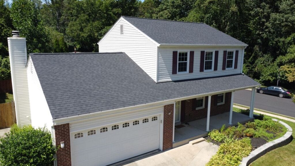 Roof Materials Lorton A two-story suburban house with white siding, red brick accents, and a dark gray shingle roof. The home features a double garage, a front porch, and is surrounded by greenery and a neatly landscaped yard.