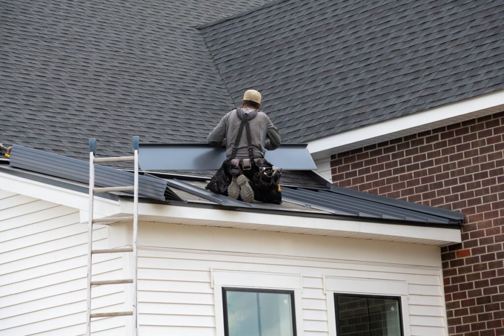 Roof Replacement Cost Falls Church A construction worker wearing a tool belt installs metal roofing panels on a house's roof. A ladder leans against the white siding, and a brick wall is visible in the background.
