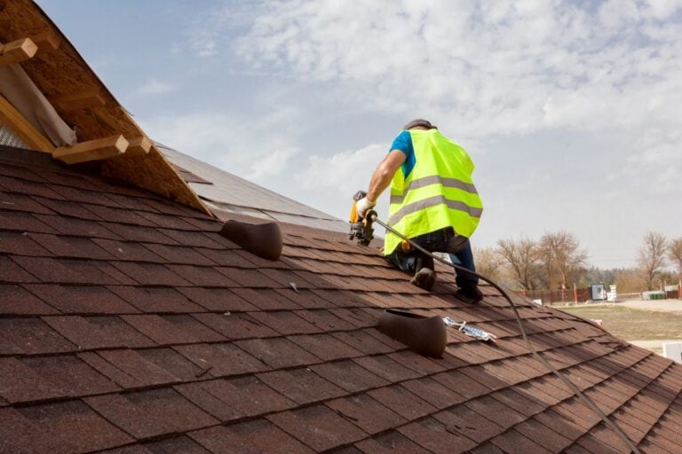 Roof Materials Lorton A construction worker in a neon safety vest installs roofing shingles on a sloped roof using a nail gun, with tools and materials nearby under a partly cloudy sky.