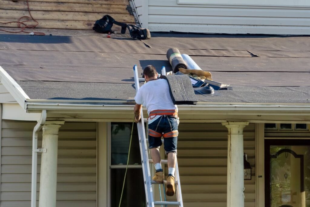 Roof Materials Lorton A man wearing a safety harness climbs a ladder to work on a house roof, which is partially covered with roofing materials and tools. The house has beige siding and white pillars on the porch.