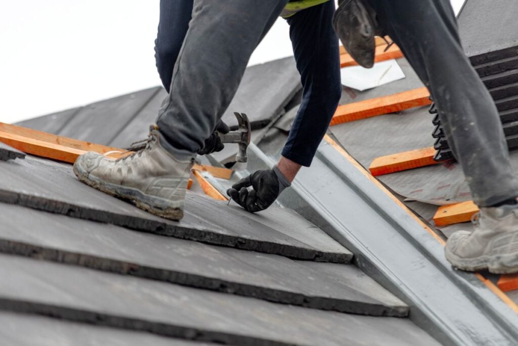 Roof Replacement Cost Falls Church worker wearing boots and gloves uses a hammer to fix roofing tiles on a sloped roof, with wooden battens and a stack of tiles visible nearby.