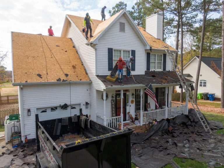 Roof Replacement Cost Falls Church Several workers are replacing the roof of a two-story house, removing old shingles and installing new ones. Debris covers the yard and porch, with a large dumpster truck parked below to collect old materials.