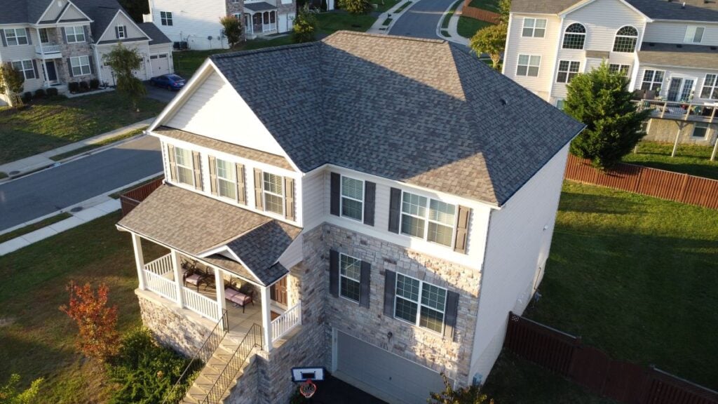 Roofing Companies In Mount Vernon VA Aerial view of a two-story suburban house with gray shingles, white siding, stone accents, a front porch, and attached garage. The house sits on a corner lot with a lawn and neighboring homes nearby.