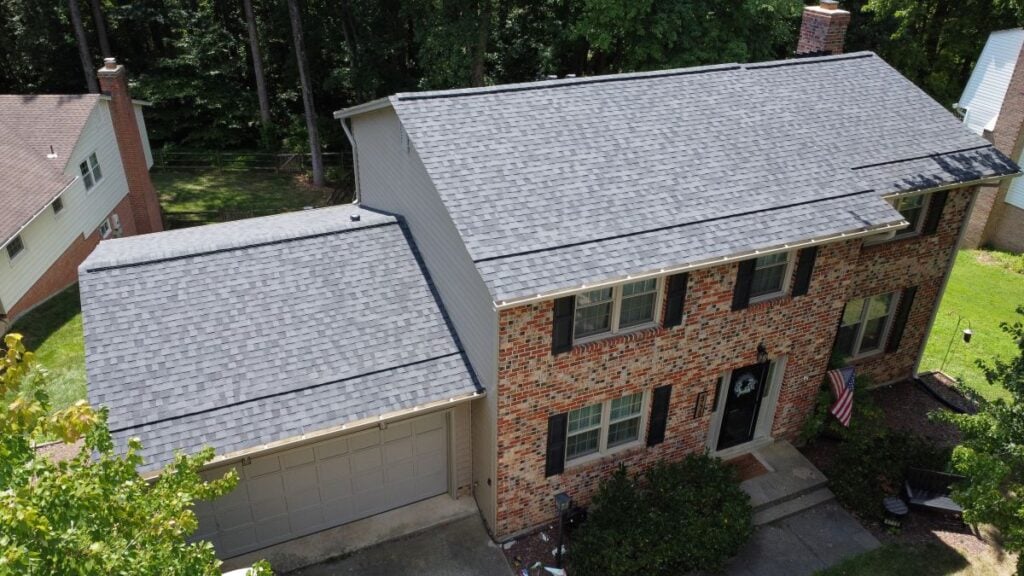 Roofing Companies In Springfield VA Aerial view of a two-story brick house with a gray shingle roof, attached garage, black shutters, and an American flag by the front door, surrounded by greenery and trees.