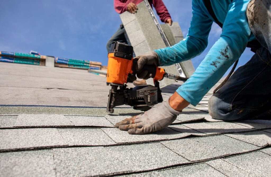 Roofing Companies In Springfield VA construction worker wearing gloves uses a nail gun to secure shingles on a roof while another worker carries shingles in the background under a clear blue sky.