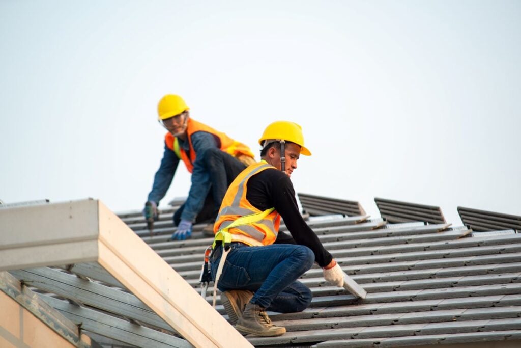Roofing Companies In Springfield VA Two construction workers wearing yellow hard hats and orange safety vests install roofing materials on a sloped roof. One is kneeling in the foreground, working with a tool, while the other is farther back, arranging tiles.