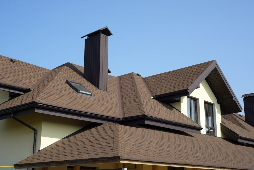 A close-up view of a house with brown shingle roofing in roofing Fairfax style, featuring multiple roof angles, a skylight, a tall chimney, and light-colored walls under a clear blue sky.