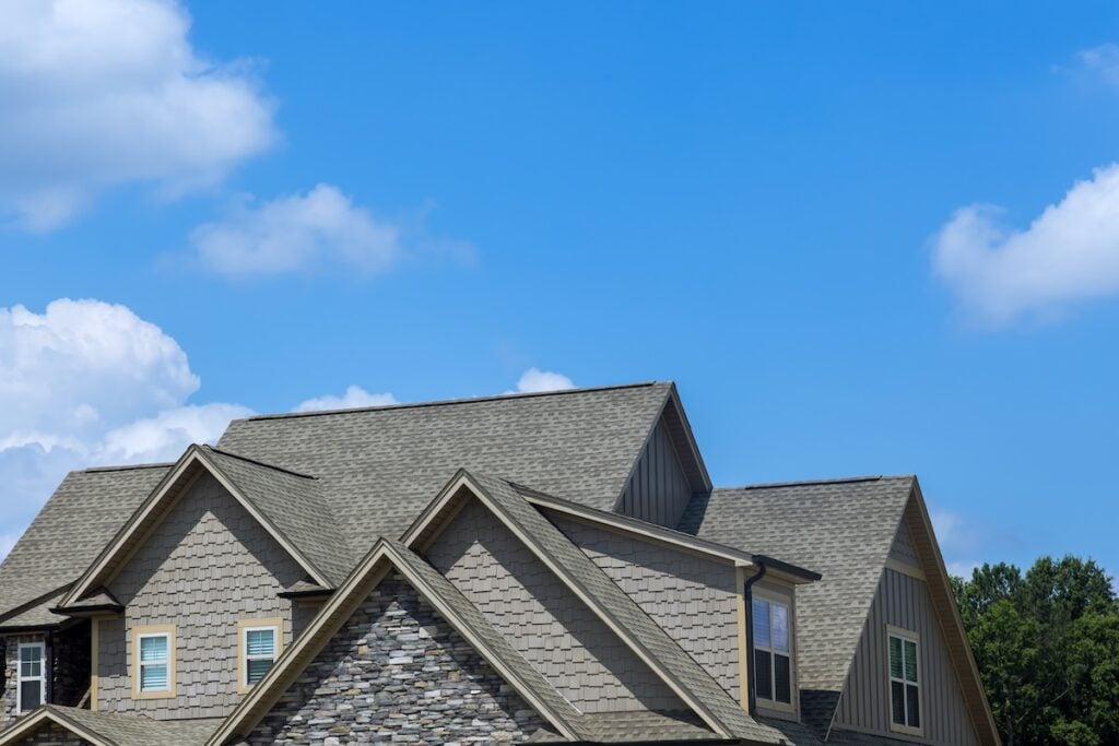 The image shows the rooftops of a modern suburban house with gray shingles and stone detailing, set against a bright blue sky and green trees—an example of quality roofing Falls Church homeowners admire.