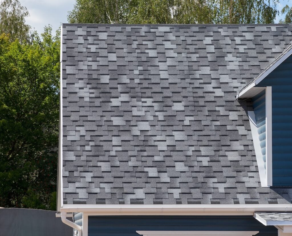 A close-up view of a house roof with gray asphalt shingles in a staggered pattern highlights popular roof materials in Falls Church. The blue house and green trees are visible in the background under a partly cloudy sky.