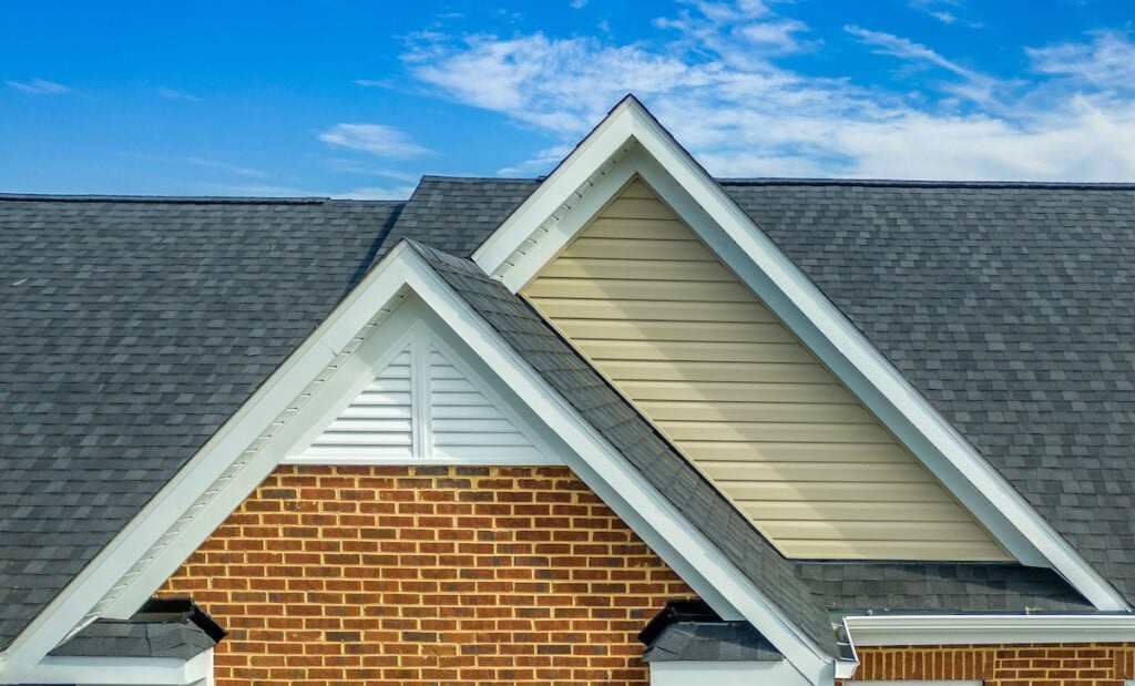 Close-up of a house roof with multiple gables, featuring gray asphalt shingles expertly installed by roofing Lorton, red brick siding, beige and white gable ends, and a bright blue sky with scattered clouds in the background.