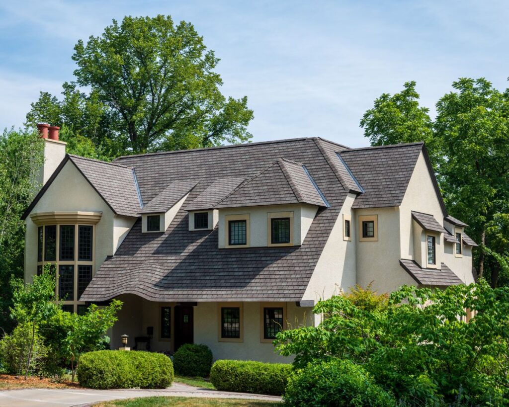 A two-story house with a large, sloped, shingle roof—expertly crafted by roofing Vienna VA—features a cream-colored exterior, many windows, and is surrounded by trees and shrubs on a sunny day.