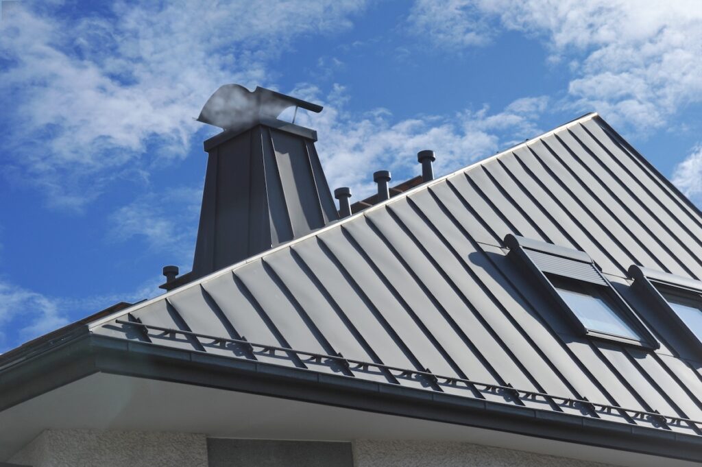 A modern house with a sleek gray metal roofing Lorton style, visible chimney, several roof vents, and a skylight window under a blue sky with scattered clouds.
