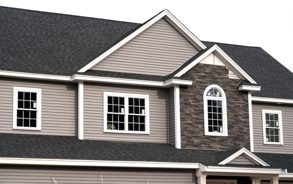 Close-up of a modern house exterior in Fairfax featuring gray siding, white-framed windows, and a stone accent around an arched window. The roofing Fairfax homeowners trust is showcased here with clean lines, dark gray finish, and multiple gables.