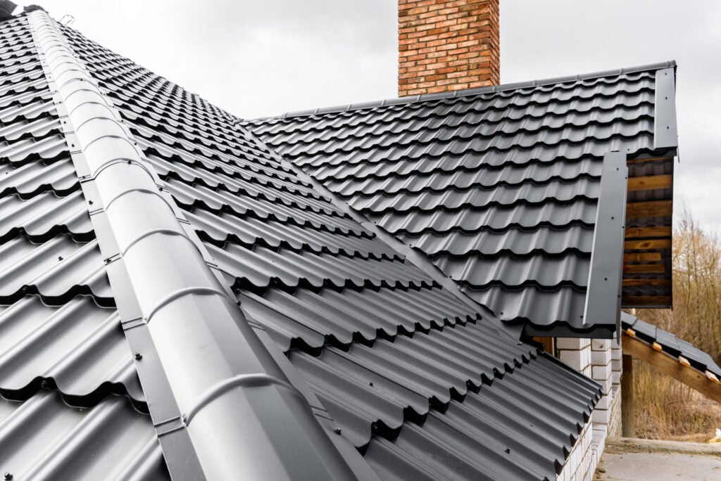 Close-up view of a house roof with black metal roofing tiles and a red brick chimney, showcasing modern, wavy textures. Perfect inspiration for roofing Falls Church homes against a dramatic, cloudy sky backdrop.