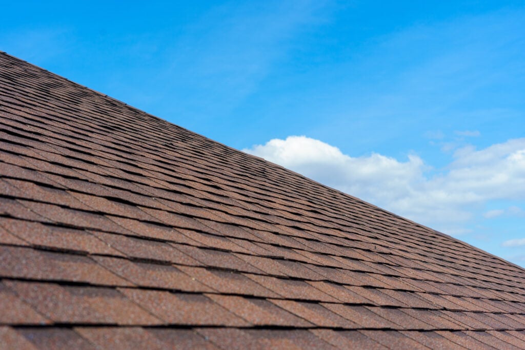 A close-up view of a sloped brown shingle roof installed by roofing Lorton, set against a bright blue sky with a few white clouds.