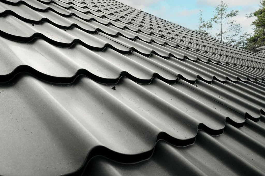 Roof Materials Lorton Close-up view of a metal roof with wavy, tile-like panels. The surface appears clean and shiny, showcasing quality Roof Materials Lorton. In the background, clouds and tree branches are visible against the sky.