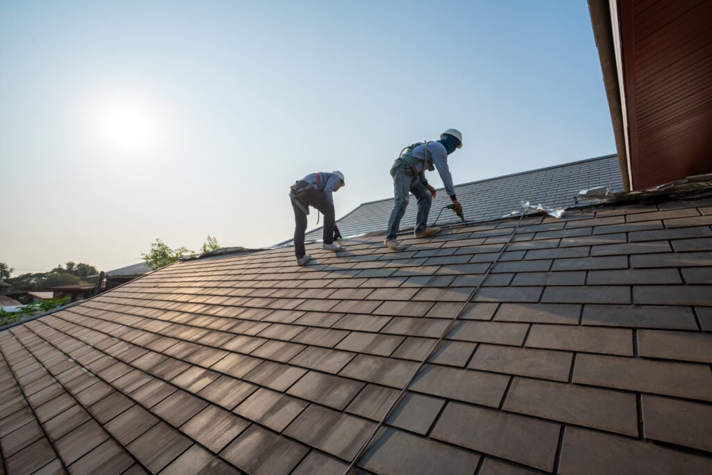 Two construction workers wearing safety harnesses install or repair shingles on a sloped roof under a clear sky with the sun shining in the background.