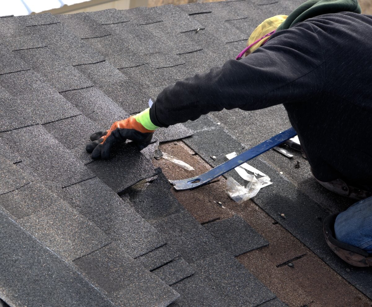 A person wearing gloves and a hat repairs shingles on a roof in Falls Church using a pry bar. Some shingles are lifted, revealing the underlayment as they work on roofing maintenance.