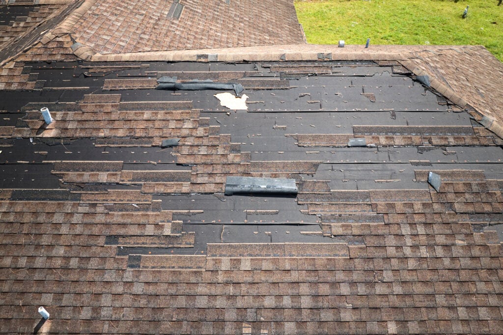 A house roof with many missing and damaged shingles, exposing the black underlayment beneath. The surrounding shingles appear worn, and some vents are visible on the roof. Grass can be seen in the background, suggesting a need for roofing Fairfax services.