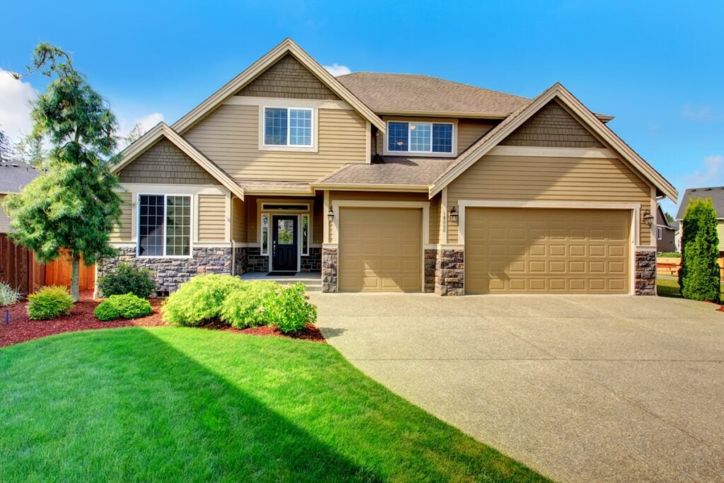 A modern two-story suburban house with beige siding, stone accents, and a three-car garage. Quality roofing Lorton enhances its curb appeal. The front yard features green grass, manicured shrubs, and a clear blue sky overhead.