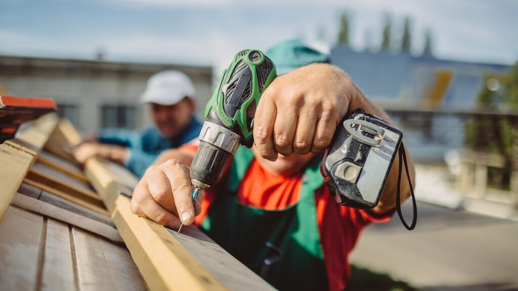 Close-up of a person using a cordless power drill to fasten a screw into a wooden board outdoors, with another person blurred in the background.