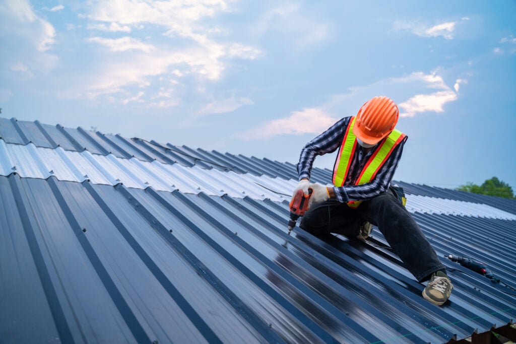 A construction worker wearing a safety vest and orange helmet uses a power drill to install or repair metal roofing panels for a roofing Fairfax project under a partly cloudy sky.