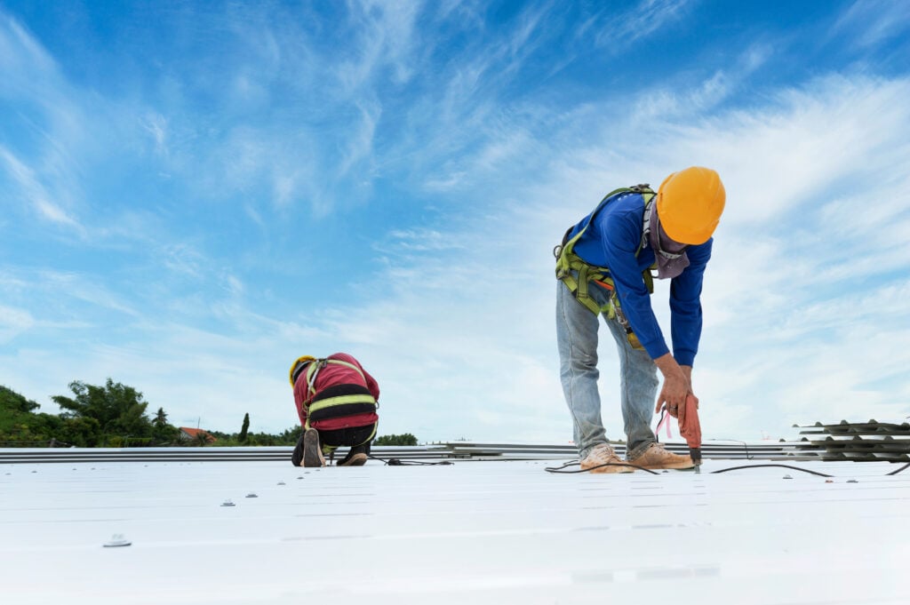 Two workers wearing safety gear and yellow helmets install or repair a white metal roof under a bright blue sky with scattered clouds. One is standing and using tools; the other is kneeling and working.