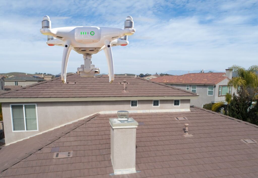 A white drone hovers above the roof of a suburban house on a clear day, capturing aerial views for roofing Vienna VA services, with neighboring homes and a blue sky in the background.